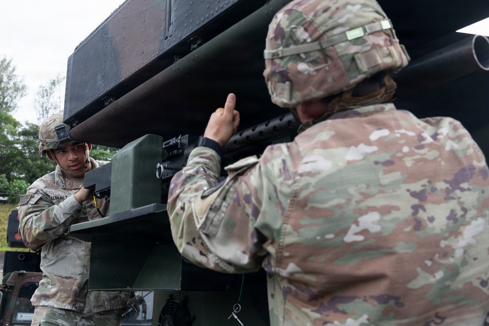 Two U.S. Army soldiers in Operational Camouflage Pattern (OCP) uniforms are working with a large military weapons system, likely an M270 Multiple Launch Rocket System (MLRS) or similar artillery platform. One soldier, whose name tape reads 'ALVAREZ,' is positioned on the left side of the launcher, while a second soldier points toward the launch tubes. The setting appears to be an outdoor training or operational environment with overcast skies.