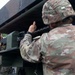 Two U.S. Army soldiers in Operational Camouflage Pattern (OCP) uniforms are working with a large military weapons system, likely an M270 Multiple Launch Rocket System (MLRS) or similar artillery platform. One soldier, whose name tape reads 'ALVAREZ,' is positioned on the left side of the launcher, while a second soldier points toward the launch tubes. The setting appears to be an outdoor training or operational environment with overcast skies.