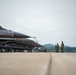 A row of F-16 Fighting Falcon jet fighters is lined up on a military airfield tarmac, armed with visible munitions under their wings. Several personnel in camouflage uniforms are standing and walking alongside the aircraft. The overcast sky and green hills in the background suggest a temperate geographic location.