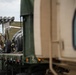 Two soldiers in camouflage uniforms are securing military equipment on a flatbed truck. The equipment appears to be a wheeled military trailer or artillery piece being loaded or tied down for transport. The scene takes place outdoors, likely on a military installation or staging area.