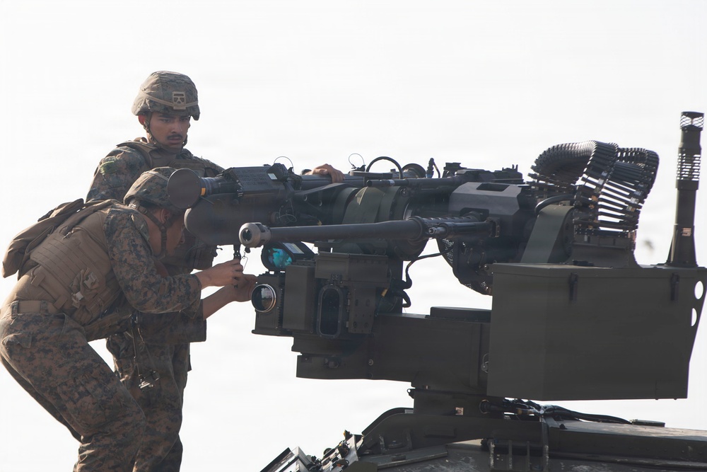 Two military personnel in camouflage uniforms and tactical gear are working on a vehicle-mounted weapon system. They appear to be conducting maintenance, loading, or configuration of what appears to be a remote weapon station or heavy machine gun mount with an ammunition belt visible. The setting appears to be an outdoor military environment with an overcast sky.