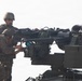 Two military personnel in camouflage uniforms and tactical gear are working on a vehicle-mounted weapon system. They appear to be conducting maintenance, loading, or configuration of what appears to be a remote weapon station or heavy machine gun mount with an ammunition belt visible. The setting appears to be an outdoor military environment with an overcast sky.