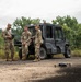Three military personnel in US Army MultiCam/OCP camouflage uniforms are gathered near a Kawasaki Mule diesel 4x4 utility vehicle on a gravel surface. One soldier is holding what appears to be a drone controller or tablet device, while a small quadcopter drone is visible on the ground nearby. The setting appears to be a training area or field exercise location surrounded by dense green vegetation.