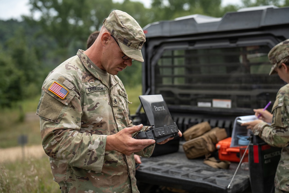 A U.S. Army soldier in MultiCam/OCP camouflage uniform is examining a Parrot brand drone controller/ground control station in an outdoor field setting. A second soldier is visible in the background taking notes near a military utility vehicle loaded with tactical gear. The scene appears to depict a drone operations or UAS (Unmanned Aerial Systems) training or evaluation exercise.