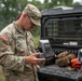 A U.S. Army soldier in MultiCam/OCP camouflage uniform is examining a Parrot brand drone controller/ground control station in an outdoor field setting. A second soldier is visible in the background taking notes near a military utility vehicle loaded with tactical gear. The scene appears to depict a drone operations or UAS (Unmanned Aerial Systems) training or evaluation exercise.