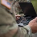 A U.S. military soldier in MultiCam/OCP camouflage uniform is operating what appears to be a drone remote control unit with an integrated screen and joystick controls. The device features a foldable display hood and touchscreen interface, consistent with modern unmanned aerial vehicle (UAV) ground control equipment. The image is a close-up shot focusing on the hands and controller, with the soldier's American flag patch visible on the left shoulder.