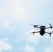 A black quadcopter drone is captured mid-flight against a pale blue, partly cloudy sky. The drone is positioned in the right-center of the frame, with its four propellers visibly spinning, indicated by motion blur. The image appears to be a professional or high-quality photograph taken from a low angle looking upward.