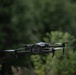 A dark-colored quadcopter drone is captured mid-flight against a blurred green foliage background. The drone appears to be a consumer or prosumer model, likely a DJI Mavic series, with four spinning propellers and a camera/sensor module mounted on its body. The image uses a shallow depth of field, keeping the drone in sharp focus while the background trees are heavily blurred.