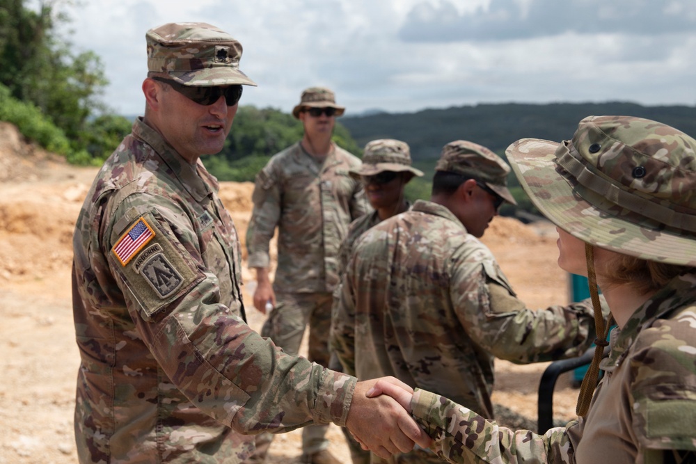 A group of U.S. Army soldiers in Operational Camouflage Pattern (OCP) uniforms are gathered at what appears to be an outdoor training or field site with sandy terrain. The soldier on the left, wearing an Airborne tab and U.S. flag patch, is shaking hands with another soldier wearing a boonie hat. Several other soldiers are visible in the background.
