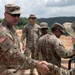 A group of U.S. Army soldiers in Operational Camouflage Pattern (OCP) uniforms are gathered at what appears to be an outdoor training or field site with sandy terrain. The soldier on the left, wearing an Airborne tab and U.S. flag patch, is shaking hands with another soldier wearing a boonie hat. Several other soldiers are visible in the background.