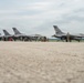 A flight line at a military airbase showing multiple F-16 Fighting Falcon jets parked in a row, with ground crew personnel performing maintenance and pre-flight checks. The aircraft display tail markings indicating they belong to the South Dakota Air National Guard. One aircraft on the far right appears to be taxiing or positioned separately from the main group.