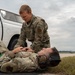 Two military personnel in OCP (Operational Camouflage Pattern) uniforms are engaged in what appears to be a tactical combat casualty care (TCCC) or first aid training exercise on a flight line or airfield. One service member is lying on the ground while the other kneels over them, appearing to assess or treat a simulated injury. The setting includes a white pickup truck and what appears to be an airfield tarmac in the background.