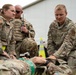 Three U.S. Air Force personnel in OCP (Operational Camouflage Pattern) uniforms are conducting what appears to be a tactical combat casualty care (TCCC) training exercise outdoors. Two personnel are kneeling over a simulated casualty on the ground, with one applying or demonstrating a medical technique while the other observes. A third service member stands behind holding a two-way radio, likely supervising or coordinating the exercise.