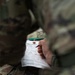 A person in military camouflage (OCP/MultiCam pattern) is filling out a TCCC (Tactical Combat Casualty Care) card, also known as a DD Form 1380 or Combat Casualty Card. The card is attached via a metal grommet and string, consistent with standard field medical documentation procedures. This appears to be a training exercise or real casualty documentation scenario.