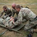 Three military personnel in camouflage uniforms are performing a tactical combat casualty care (TCCC) exercise on a fourth individual lying on a green field litter/stretcher. The scene appears to be an outdoor training exercise, with one female and two male soldiers attending to the simulated casualty. A blue and white boundary rope is visible in the background, suggesting a designated training area.