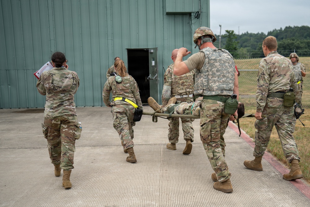 Military personnel in camouflage uniforms are carrying a casualty on a litter/stretcher toward the entrance of a green metal building. The scene appears to be a medical evacuation drill or training exercise, with multiple soldiers participating in coordinated roles. One soldier carries documents, another wears hearing protection, and at least one wears a tactical vest.