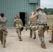 Military personnel in camouflage uniforms are carrying a casualty on a litter/stretcher toward the entrance of a green metal building. The scene appears to be a medical evacuation drill or training exercise, with multiple soldiers participating in coordinated roles. One soldier carries documents, another wears hearing protection, and at least one wears a tactical vest.