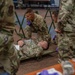 Military medical personnel in OCP (Operational Camouflage Pattern) uniforms are conducting a medical training exercise. A simulated casualty is lying on a military litter/stretcher on the floor with medical tubing attached, while at least two medics wearing purple nitrile gloves attend to him. One personnel has a 'CCATT' (Critical Care Air Transport Team) patch visible on their uniform.