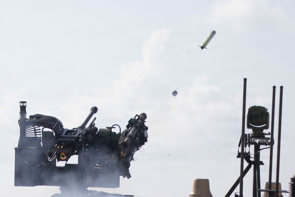 The image captures a military weapons system in the process of firing a missile or rocket, which is visibly airborne in the upper right portion of the frame. Smoke and debris are visible around the launcher, indicating a recent or ongoing firing event. A secondary sensor or radar unit is visible on the right side of the image.