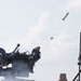 The image captures a military weapons system in the process of firing a missile or rocket, which is visibly airborne in the upper right portion of the frame. Smoke and debris are visible around the launcher, indicating a recent or ongoing firing event. A secondary sensor or radar unit is visible on the right side of the image.