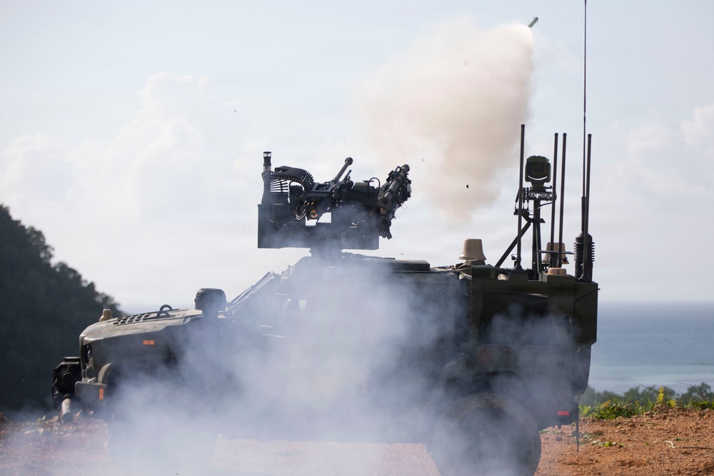 A military armored vehicle, consistent with a Stryker-type wheeled APC, is actively firing a weapon system mounted on its turret. A projectile or missile is visible leaving the launcher with a large smoke plume billowing around the vehicle. The scene appears to be an outdoor live-fire exercise or demonstration near a coastal or lakeside environment.