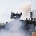 A military armored vehicle, consistent with a Stryker-type wheeled APC, is actively firing a weapon system mounted on its turret. A projectile or missile is visible leaving the launcher with a large smoke plume billowing around the vehicle. The scene appears to be an outdoor live-fire exercise or demonstration near a coastal or lakeside environment.