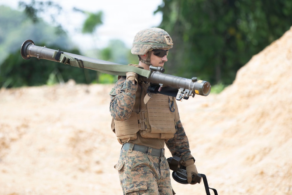A male soldier in US Marine Corps MARPAT digital camouflage uniform carries a shoulder-launched rocket system (consistent with an AT4 or similar anti-armor weapon) resting on his right shoulder. He is equipped with full combat gear including a plate carrier vest, ballistic helmet, and protective eyewear. The setting appears to be a training range or sandy outdoor environment with vegetation visible in the background.