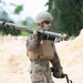 A male soldier in US Marine Corps MARPAT digital camouflage uniform carries a shoulder-launched rocket system (consistent with an AT4 or similar anti-armor weapon) resting on his right shoulder. He is equipped with full combat gear including a plate carrier vest, ballistic helmet, and protective eyewear. The setting appears to be a training range or sandy outdoor environment with vegetation visible in the background.
