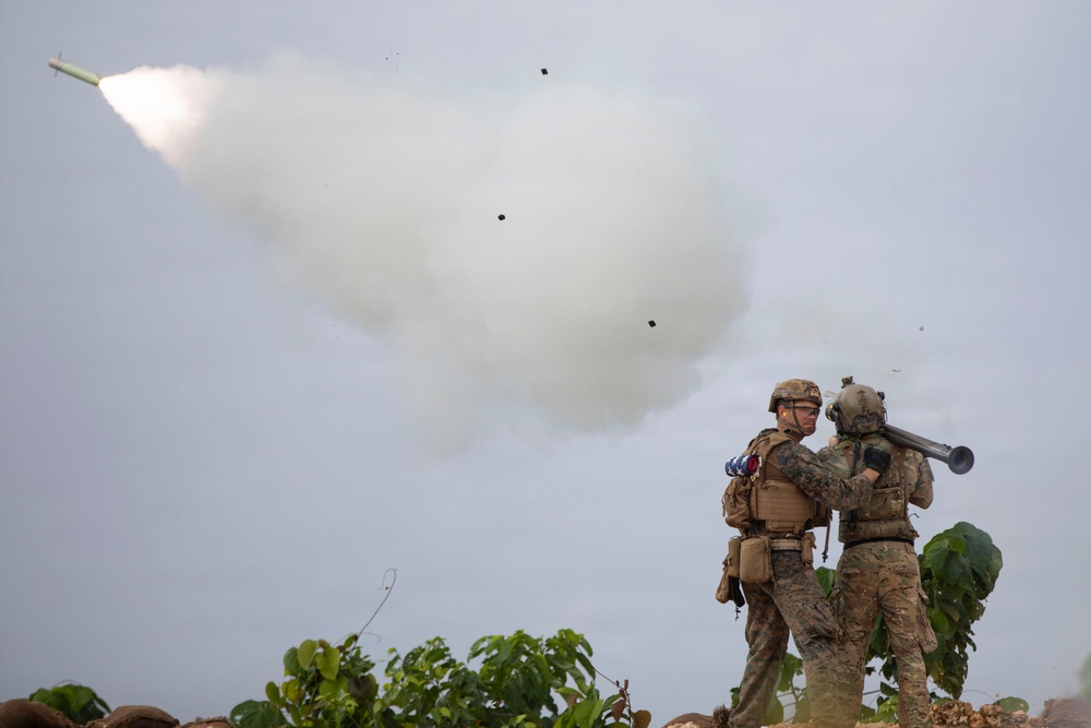 Two military personnel in full combat gear are positioned on elevated terrain as a rocket or missile is launched into the sky, leaving a large white smoke trail. One soldier appears to be holding a launch tube (likely an AT4 or similar shoulder-fired weapon) while the other observes. Debris and ejected components are visible in the air following the launch.