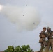 Two military personnel in full combat gear are positioned on elevated terrain as a rocket or missile is launched into the sky, leaving a large white smoke trail. One soldier appears to be holding a launch tube (likely an AT4 or similar shoulder-fired weapon) while the other observes. Debris and ejected components are visible in the air following the launch.