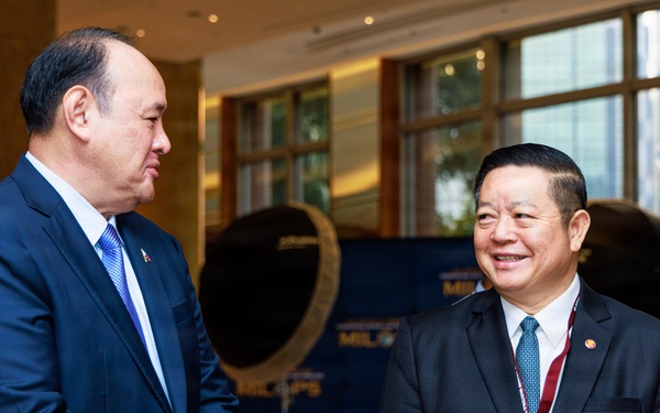 Two men in dark business suits are engaged in conversation at what appears to be a formal conference or official event. Both wear ties and lapel pins, suggesting they may be officials or delegates. The setting includes branded backdrop signage partially reading 'MIL...PS' and a decorative gong in the background.