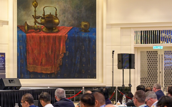 A formal conference or symposium is taking place in an elegant ballroom or hotel venue. A speaker in a dark suit addresses a large audience seated at round tables, many of whom appear to be military officers and officials in uniform. The podium bears the 'MILOPS' branding, indicating this is a military law and operations strategic engagement conference.