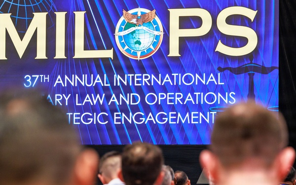 A speaker addresses a large audience at the 37th Annual MILOPS (Military Law and Operations Strategic Engagement) conference. The event appears to be a formal international military law symposium with a diverse audience seated in rows facing a stage. A large branded backdrop and podium with the MILOPS logo are prominently displayed.