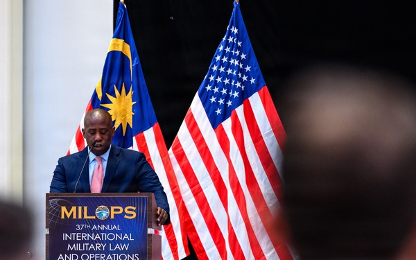 A speaker addresses an audience at the 37th Annual MILOPS (International Military Law and Operations Strategic Engagement) conference, held in Kuala Lumpur, Malaysia. The man stands at a podium flanked by the flags of Malaysia and the United States, indicating a bilateral or international military legal forum. Audience members are visible in the foreground, slightly out of focus.