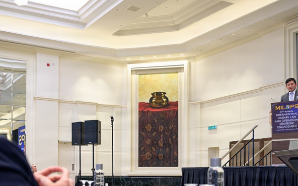 A formal conference or symposium is taking place in an elegant ballroom-style venue. A speaker stands at a podium bearing the 'MILOPS' logo, identified as the International Military Law and Operations Strategic Engagement event. Malaysian and American flags are displayed on stage, indicating a bilateral or international military engagement context.