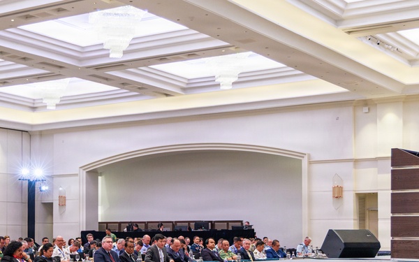 A formal conference or summit is taking place in a large ballroom or convention hall. A speaker in a dark suit stands at a wooden podium on a raised stage, addressing a large audience seated at round tables. The audience includes individuals in both military uniforms and civilian attire, suggesting a high-level defense or governmental gathering.