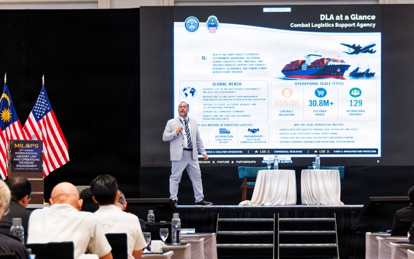 A presenter is delivering a briefing on the DLA (Defense Logistics Agency) at what appears to be the MILOPS International Military and Operations Strategic Engagement conference. The slide displayed is titled 'DLA at a Glance - Combat Logistics Support Agency' and highlights operational scale metrics including $55-4B in contract obligations, 30.8M+ customer orders, and 129 international partners. The event appears to be held in Malaysia, as indicated by the Malaysian flag visible on stage alongside the US flag.