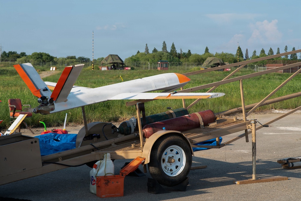A fixed-wing unmanned aerial vehicle (UAV) with white and orange markings is mounted on a wheeled ground support trailer, likely at a military or testing airfield. The trailer appears to be a launch or transport platform equipped with cylindrical objects that may be rocket-assisted launch boosters. The setting appears to be an outdoor operational area with grass, trees, and fencing visible in the background.