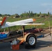 A fixed-wing unmanned aerial vehicle (UAV) with white and orange markings is mounted on a wheeled ground support trailer, likely at a military or testing airfield. The trailer appears to be a launch or transport platform equipped with cylindrical objects that may be rocket-assisted launch boosters. The setting appears to be an outdoor operational area with grass, trees, and fencing visible in the background.