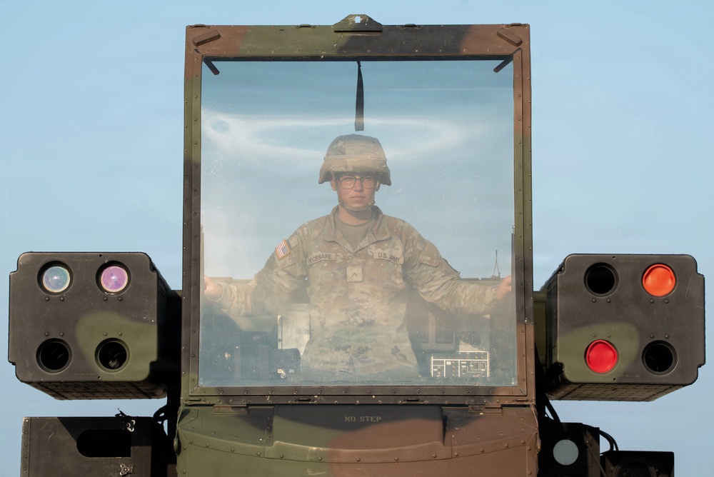 A U.S. Army soldier wearing a combat helmet and glasses is visible through the windshield of a military vehicle, likely a Humvee or similar tactical platform. The vehicle features sensor/optic pods mounted on both sides of the windshield frame, and a 'NO STEP' warning label is visible on the vehicle body. The image is taken from a front-facing perspective against a clear blue sky background.