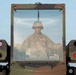 A U.S. Army soldier wearing a combat helmet and glasses is visible through the windshield of a military vehicle, likely a Humvee or similar tactical platform. The vehicle features sensor/optic pods mounted on both sides of the windshield frame, and a 'NO STEP' warning label is visible on the vehicle body. The image is taken from a front-facing perspective against a clear blue sky background.