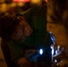A person wearing safety goggles and blue latex gloves is working on a mechanical or technical task at night, illuminated by a bright handheld flashlight. They are bent over their work, closely inspecting or manipulating an object in low-light conditions. The warm ambient background lighting contrasts with the cool blue-white light of the flashlight.