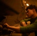 A young person with curly hair and a forearm tattoo is working intently on a large metal object in a dimly lit space. They are wearing a green long-sleeve shirt and appear to be using hand tools on a riveted metal surface. The warm, low-key lighting creates a dramatic, industrial atmosphere.