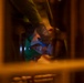 A child or young person is lying down and looking at a glowing mobile device screen in a dimly lit, warm-toned environment. The image is captured through what appears to be furniture slats or railings, creating a framed, voyeuristic perspective. The blue light from the device contrasts sharply with the surrounding amber/orange ambient lighting.
