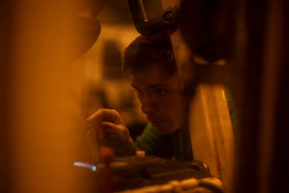 A young man is closely focused on a mechanical or technical task, viewed through a narrow gap between objects. The scene is bathed in warm amber/orange light, creating a low-light workshop or industrial environment. He appears to be manipulating a small component or tool with his hand near machinery.