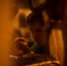 A young man is closely focused on a mechanical or technical task, viewed through a narrow gap between objects. The scene is bathed in warm amber/orange light, creating a low-light workshop or industrial environment. He appears to be manipulating a small component or tool with his hand near machinery.