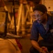 A man wearing safety goggles and a blue work uniform is crouched down in what appears to be an industrial workshop or garage. He is focused on operating or inspecting equipment in a dimly lit environment with warm orange ambient lighting. The setting suggests active mechanical or industrial work.