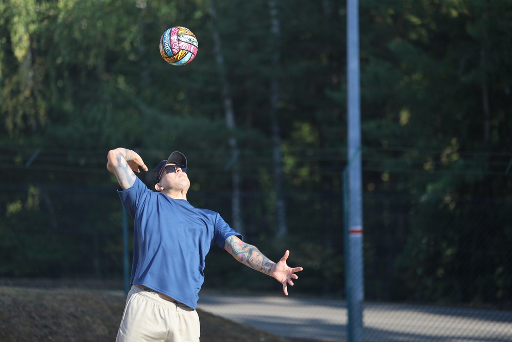 A tattooed man wearing a blue t-shirt, white shorts, a black cap, and sunglasses is playing volleyball outdoors. He appears to be in the motion of serving or setting a colorfully decorated volleyball. The scene takes place on an outdoor court surrounded by green trees and a metal fence.