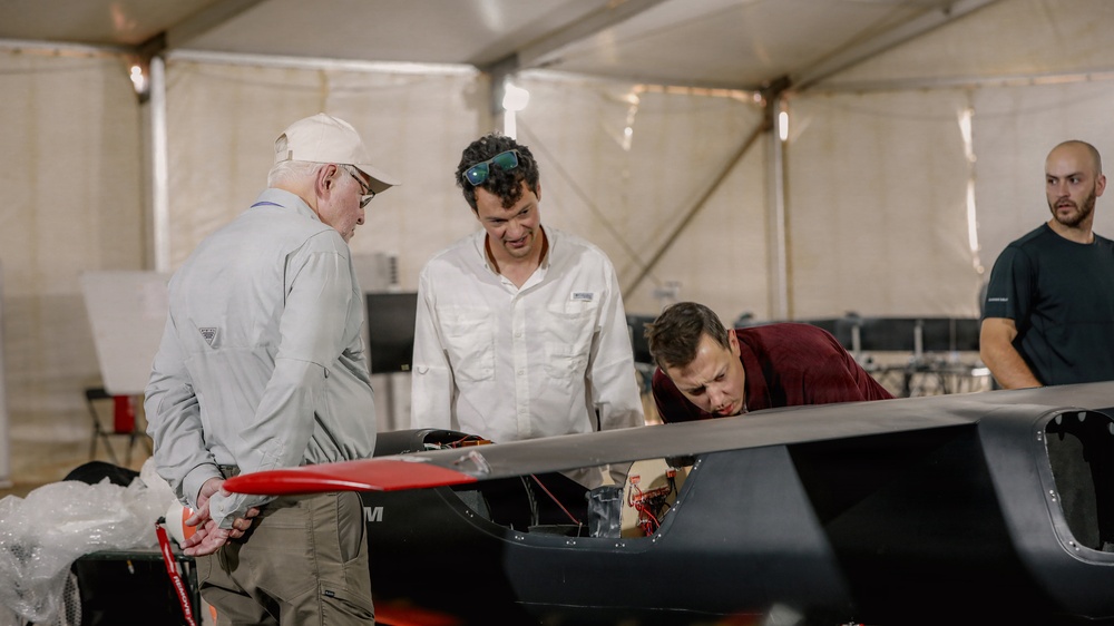 Four individuals are gathered inside a large tent or temporary structure, examining what appears to be a sleek, dark-colored aircraft or racing vehicle with red accents. Three of the men are closely inspecting the open cockpit area of the craft, while a fourth stands slightly apart in the background. The setting suggests a technical inspection, assembly, or pre-event preparation scenario.