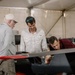 Four individuals are gathered inside a large tent or temporary structure, examining what appears to be a sleek, dark-colored aircraft or racing vehicle with red accents. Three of the men are closely inspecting the open cockpit area of the craft, while a fourth stands slightly apart in the background. The setting suggests a technical inspection, assembly, or pre-event preparation scenario.